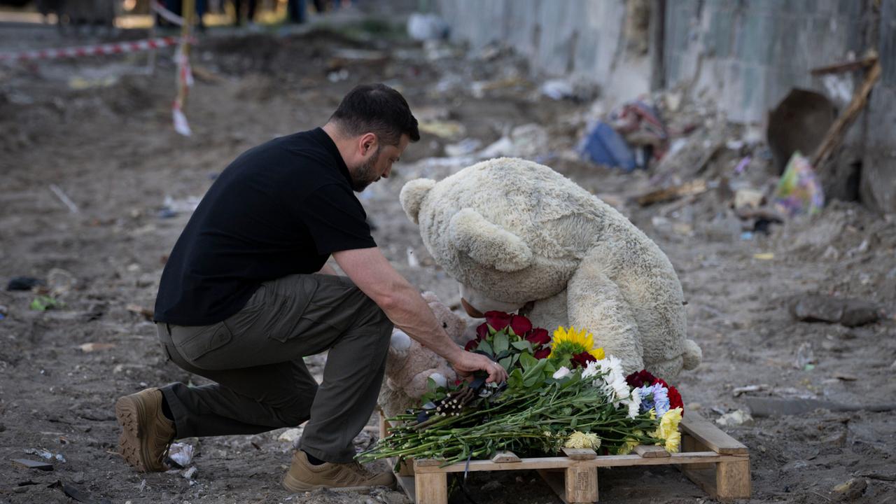 Der ukrainische Präsident Selenskyj kniet an einem Gedenkort für Todesopfer eines Bombenangriffs auf Kiew nieder und legt Blumen vor einem großen Plüschbären ab.