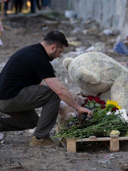 Der ukrainische Präsident Selenskyj kniet an einem Gedenkort für Todesopfer eines Bombenangriffs auf Kiew nieder und legt Blumen vor einem großen Plüschbären ab.