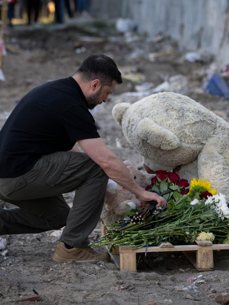 Der ukrainische Präsident Selenskyj kniet an einem Gedenkort für Todesopfer eines Bombenangriffs auf Kiew nieder und legt Blumen vor einem großen Plüschbären ab.
