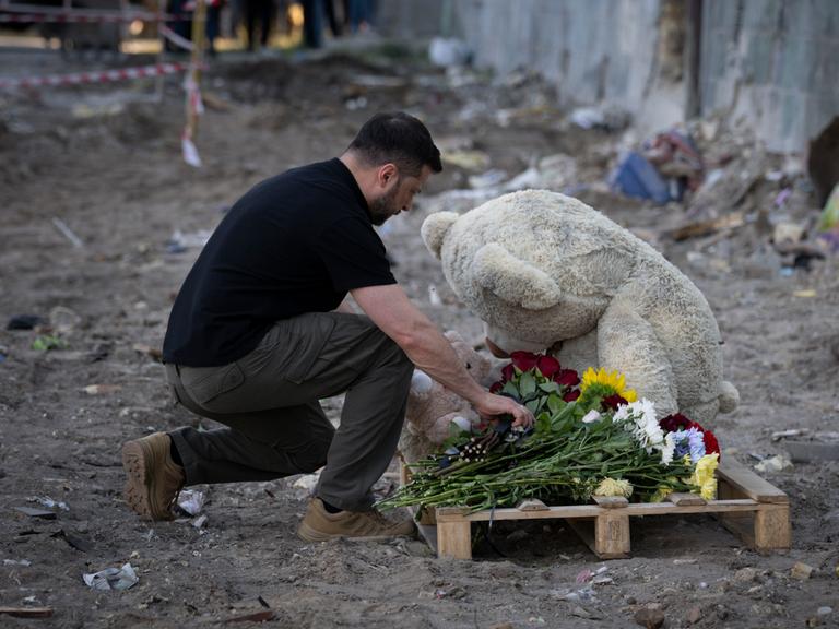 Der ukrainische Präsident Selenskyj kniet an einem Gedenkort für Todesopfer eines Bombenangriffs auf Kiew nieder und legt Blumen vor einem großen Plüschbären ab.