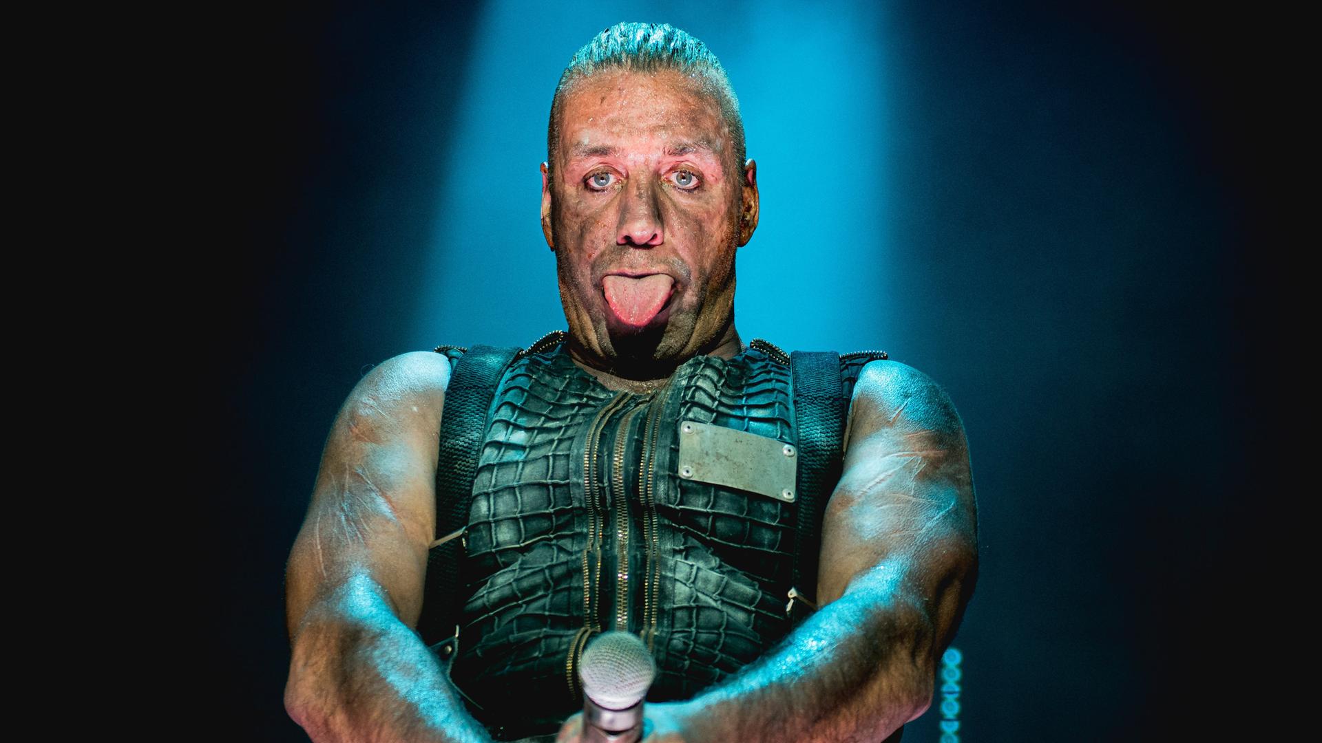 Rammstein-Frontmann Till Lindemann steht auf der Bühne und streckt die Zunge heraus. Sein Mikrofon hält er auf Hüfthöhe.