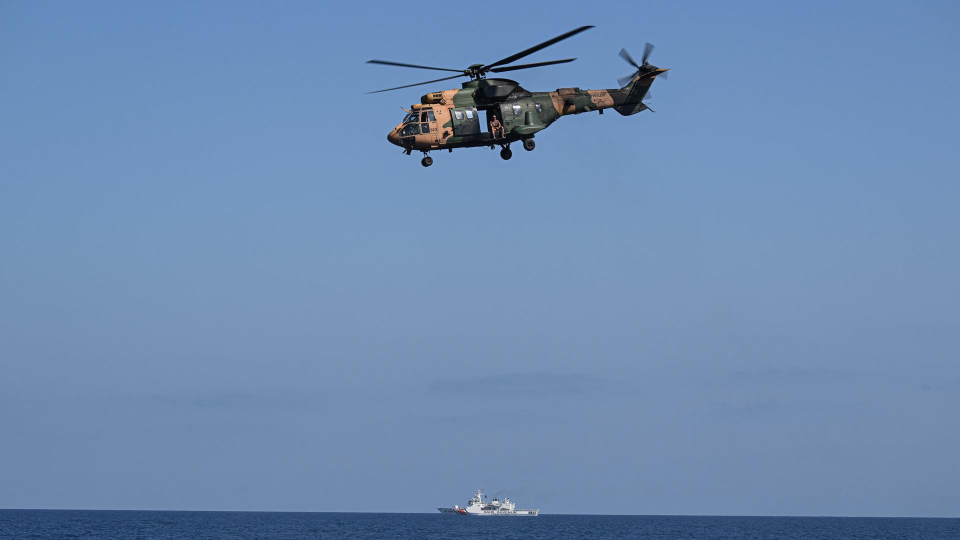 Ein Hubschrauber fliegt über dem Meer, am Horizont ist ein weißes Schiff erkennbar.