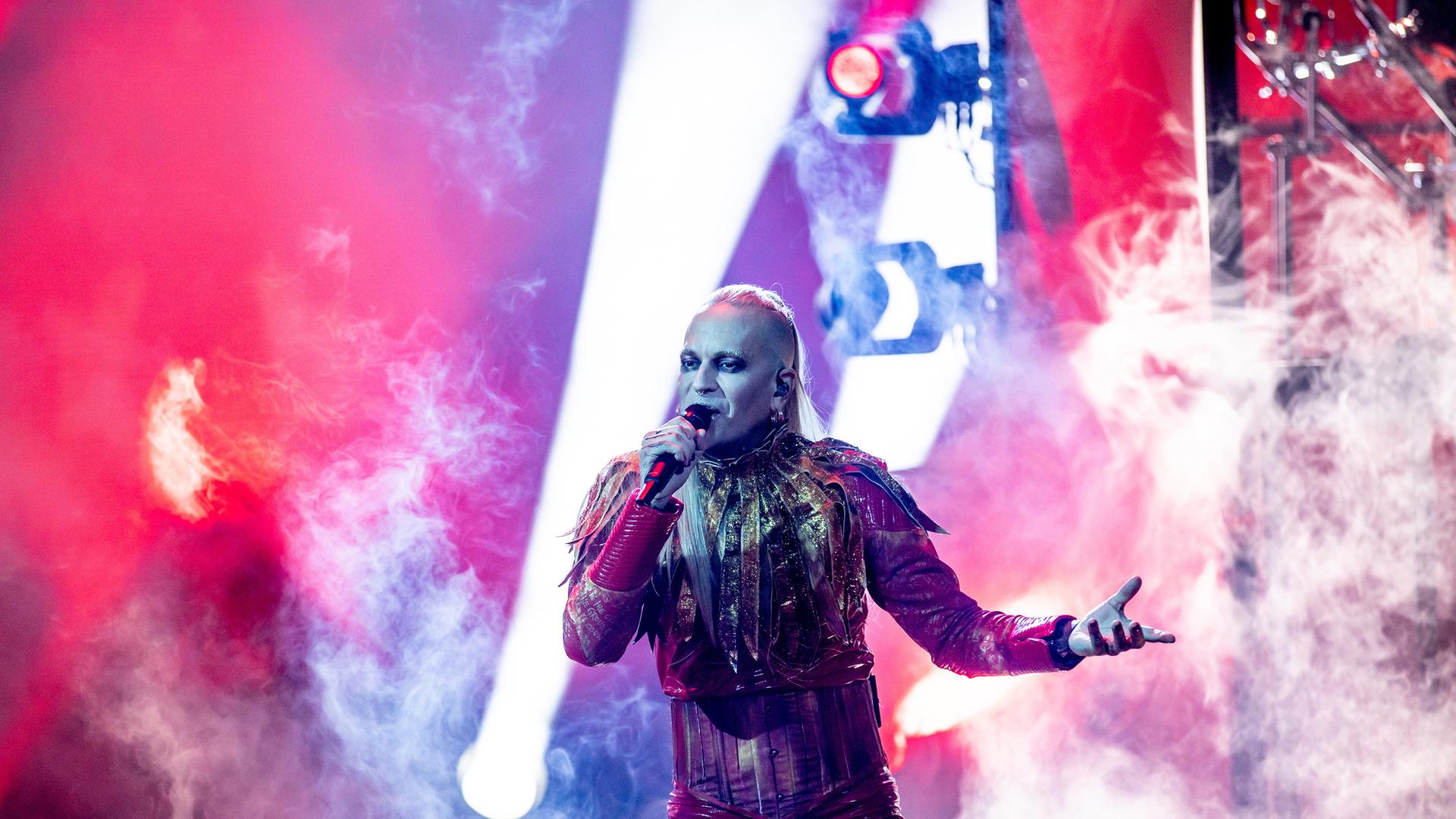 Lord of the Lost-Frontmann Chris Harms steht in seinem Kostüm, geschminkt und mit langem blondem Haar auf der Bühne und singt in ein Mikrofon.