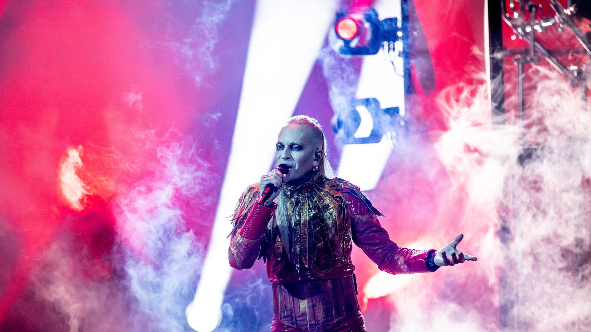 Lord of the Lost-Frontmann Chris Harms steht in seinem Kostüm, geschminkt und mit langem blondem Haar auf der Bühne und singt in ein Mikrofon. Lord of the Lost-Frontmann Chris Harms steht in seinem Kostüm, geschminkt und mit langem blondem Haar auf der Bühne und singt in ein Mikrofon.