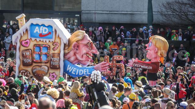 Auf dem Wagen sieht man eine Figur von Alice Weidel. Sie sieht aus wie eine Hexe und lockt zwei junge Menschen mit einem Haken-Kreuz aus Lebkuchen. in ihr Hexen-Haus.