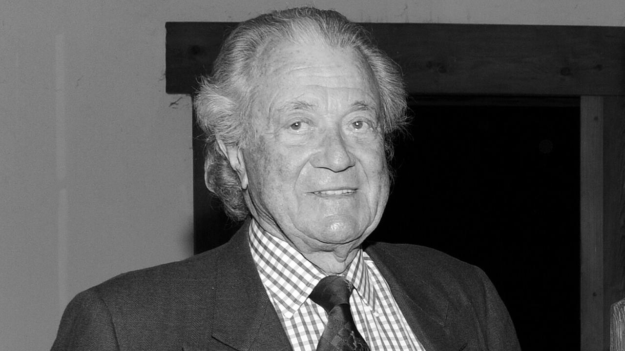 Das schwarz-weiße Bild zeigt den Oberkörper des grauhaarigen Meggle mit Anzug und Krawatte vor einer Wand. Er lächelt. 