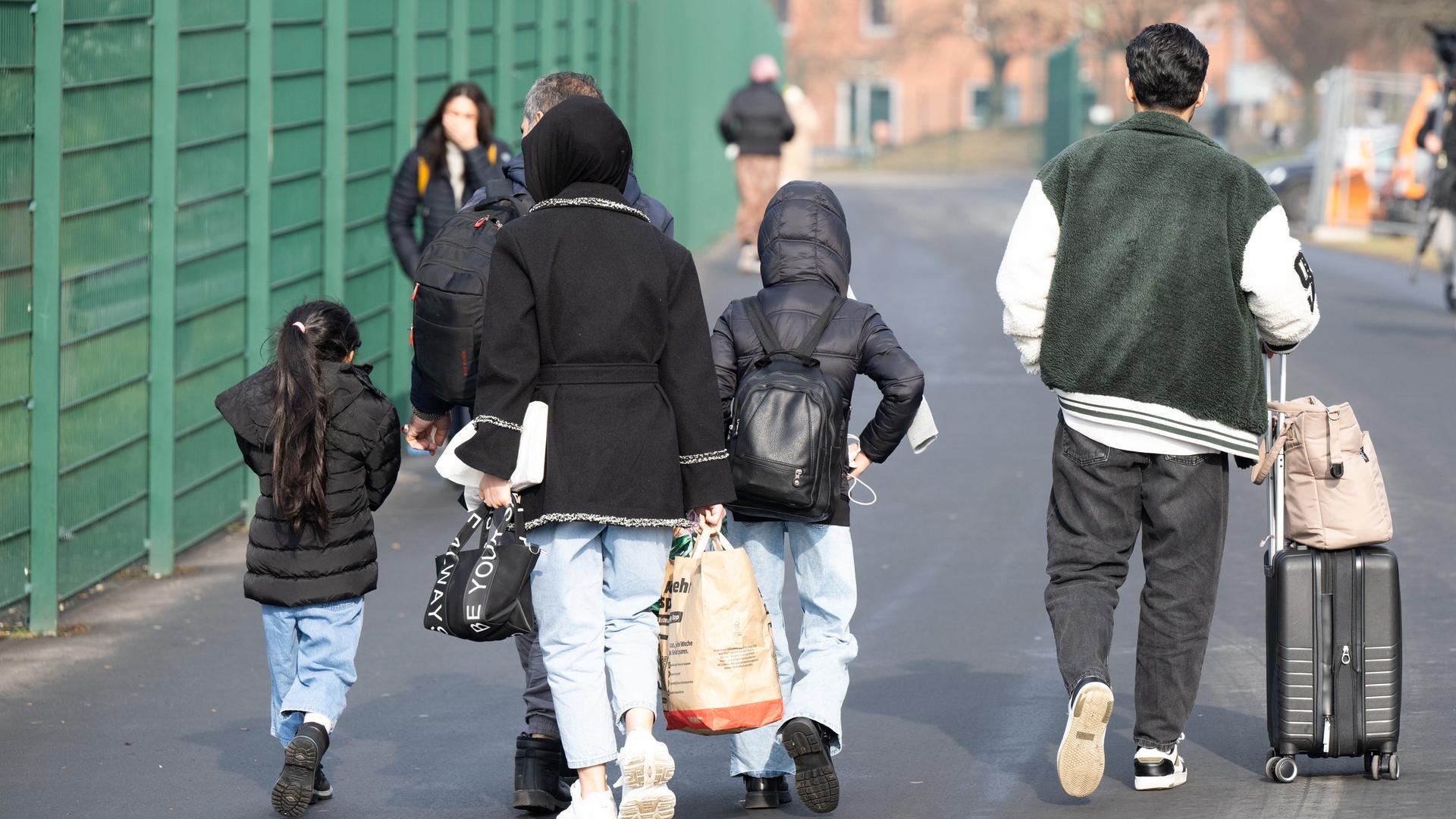 Eine Familie geht an einem Zaun vorbei. Die Menschen haben Taschen in der Hand, der Mann schiebt einen Koffer. Alle sind von hinten zu sehen. Eine Familie geht an einem Zaun vorbei. Die Menschen haben Taschen in der Hand, der Mann schiebt einen Koffer. Alle sind von hinten zu sehen.