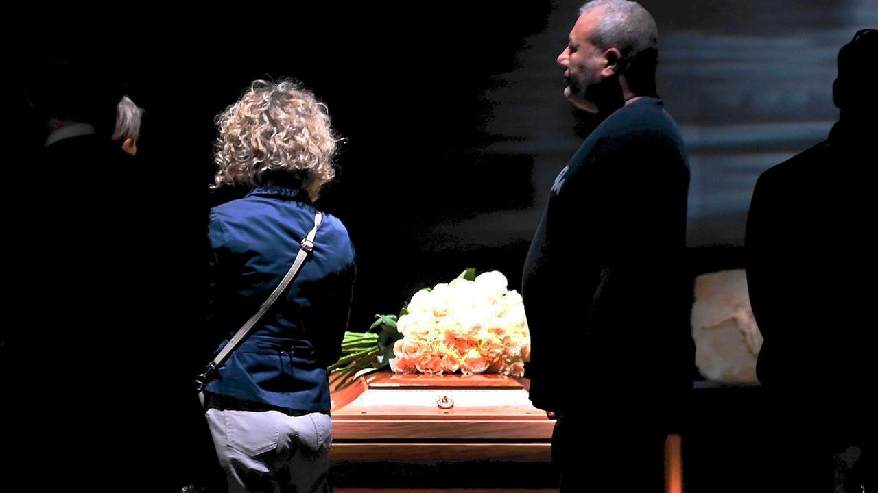 Ein Sarg mit weißen Rosen darauf ist aufgebahrt, daran gehen Menschen mit ernsten Gesichtern vorbei. 