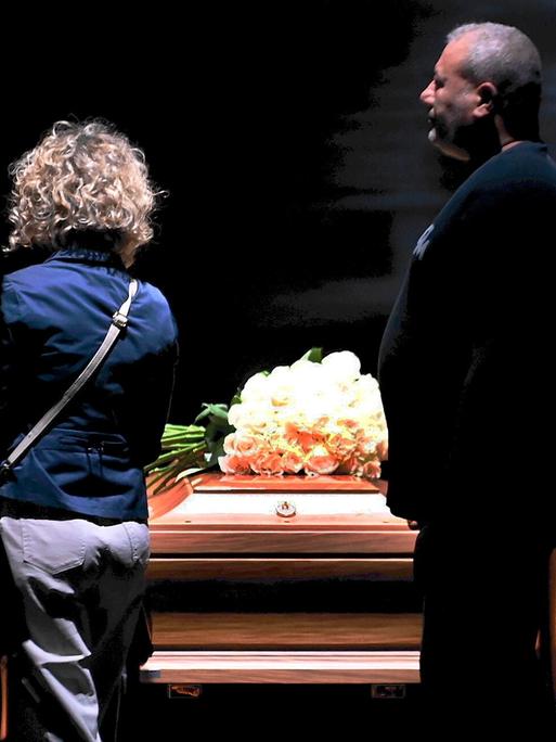 Ein Sarg mit weißen Rosen darauf ist aufgebahrt, daran gehen Menschen mit ernsten Gesichtern vorbei. 