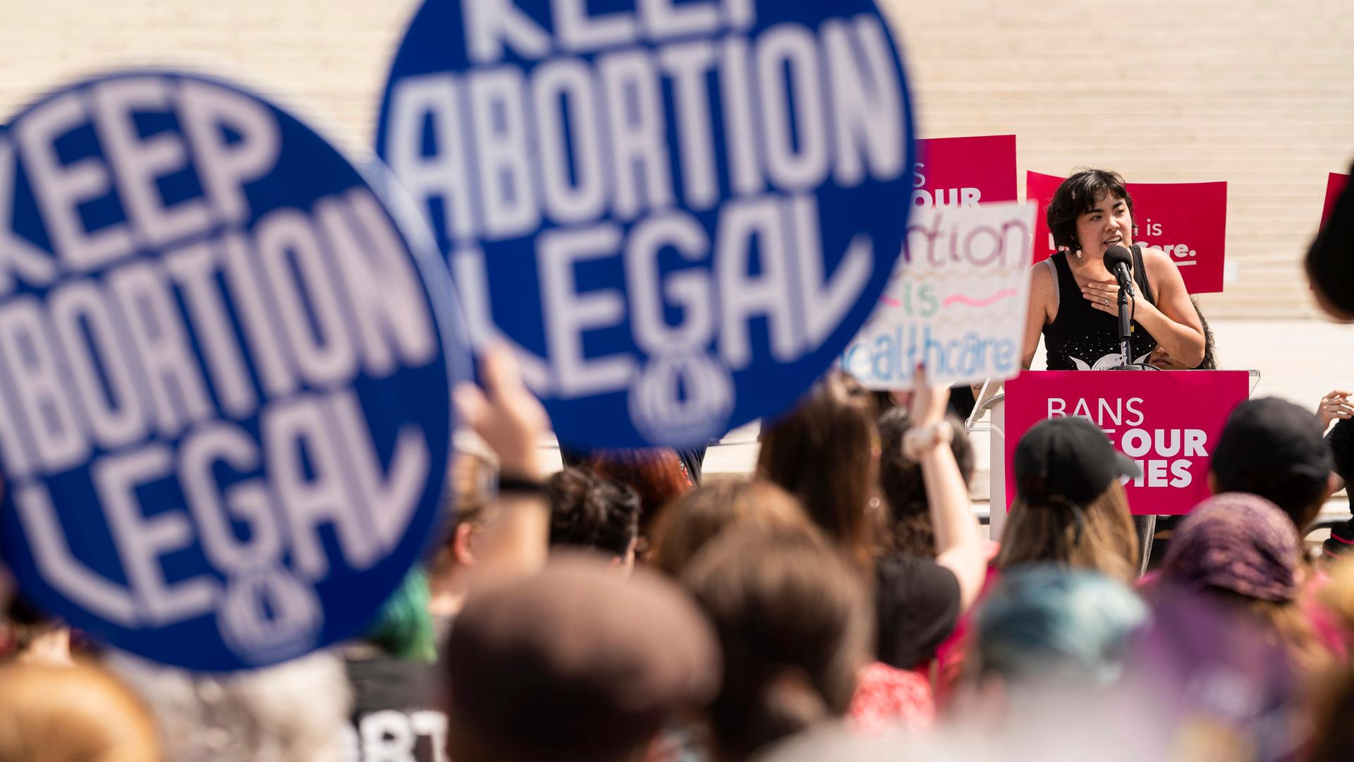 Eine Frau steht an einem Rednerpult und spricht vor Demonstrierenden. Diese halten runde, blaue Schilder in die Höhe mit der Aufschrift: "Keep Abortion Legal", was auf Deutsch soviel heißt wie "Abtreibung muss legal bleiben".