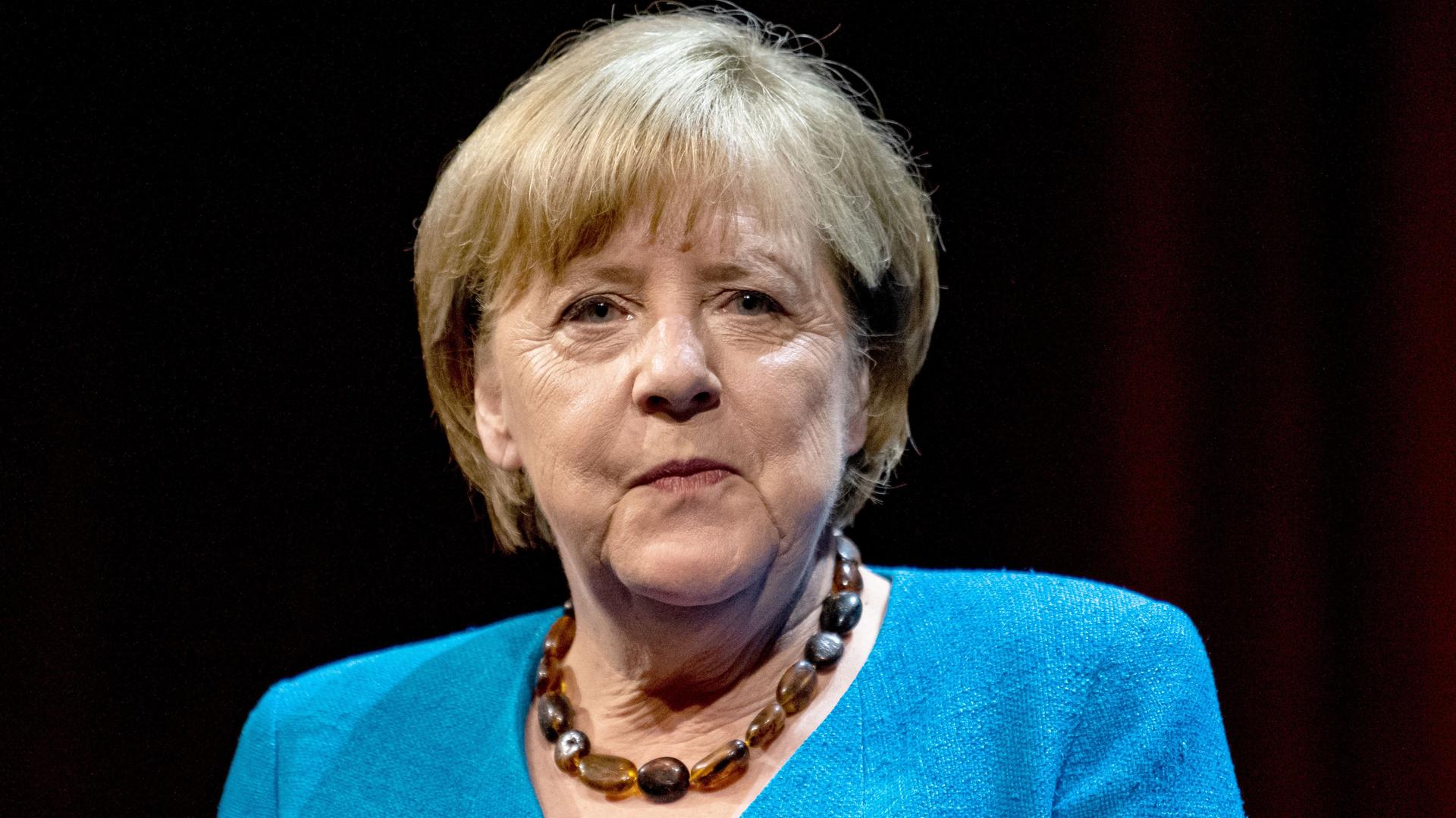 Porträt Angela Merkel: Sie trägt ein mittelblaues Kostüm und eine Halskette mit bernsteinfarbenen großen Steinen. Sie schaut ernst.