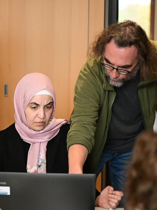 Eine Frau mit Kopftuch sitzt an einem Computer. Rechts beugt sich ein Mann zu ihr und erklärt ihr etwas am Monitor. Links steht ein weiterer Mann mit Kamera in der Hand.