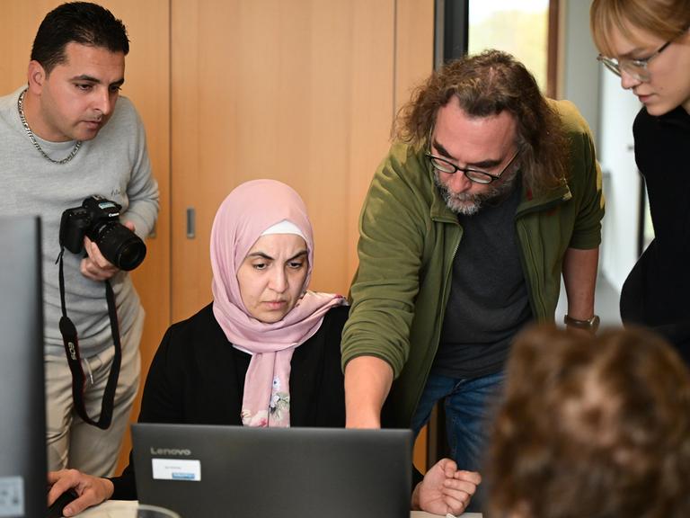 Eine Frau mit Kopftuch sitzt an einem Computer. Rechts beugt sich ein Mann zu ihr und erklärt ihr etwas am Monitor. Links steht ein weiterer Mann mit Kamera in der Hand.