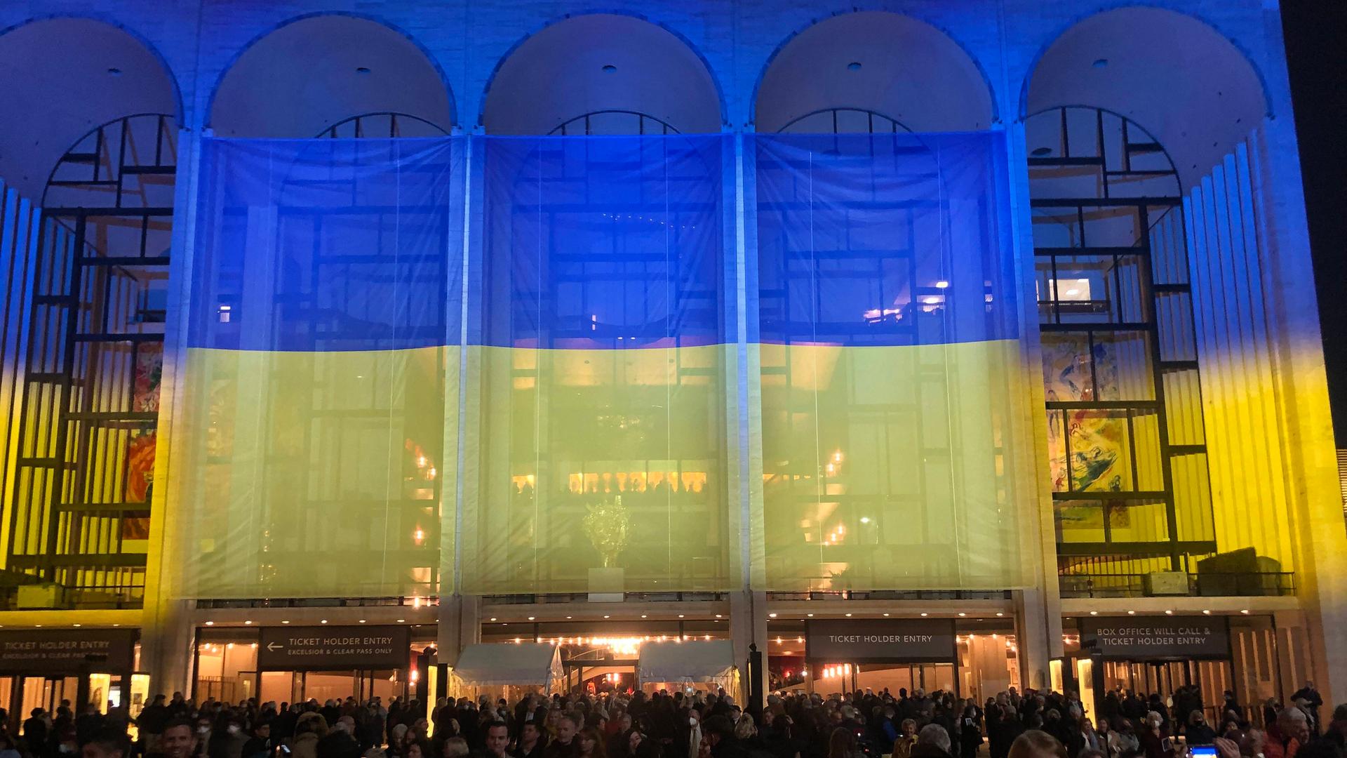 Vor den großen Eingangsbögen der MET ist eine große blau-gelbe Fahne gespannt, auf die viele Menschen zulaufen.