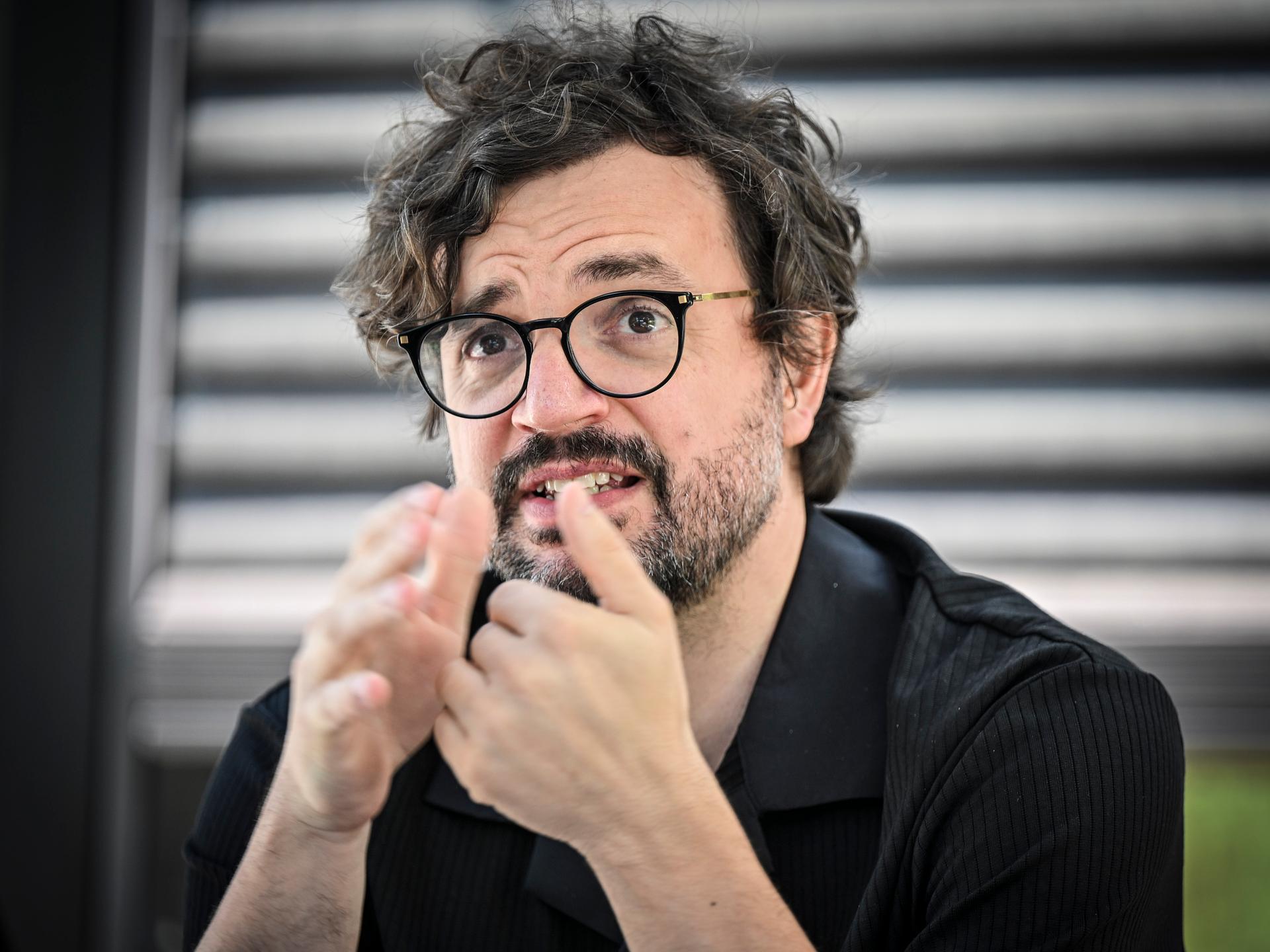 Julien Chavaz, Generalintendant des Theaters Magdeburg, während eines Interviews. Er trägt eine schwarze Brille, ein schwarzes Hemd, Bart und gestikuliert mit den Händen. Der Hintergrund ist verschwommen. Julien Chavaz, Generalintendant des Theaters Magdeburg, während eines Interviews. Er trägt eine schwarze Brille, ein schwarzes Hemd, Bart und gestikuliert mit den Händen. Der Hintergrund ist verschwommen.