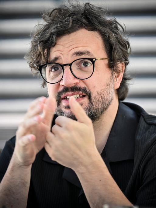 Julien Chavaz, Generalintendant des Theaters Magdeburg, während eines Interviews. Er trägt eine schwarze Brille, ein schwarzes Hemd, Bart und gestikuliert mit den Händen. Der Hintergrund ist verschwommen.