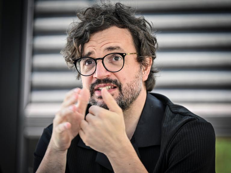 Julien Chavaz, Generalintendant des Theaters Magdeburg, während eines Interviews. Er trägt eine schwarze Brille, ein schwarzes Hemd, Bart und gestikuliert mit den Händen. Der Hintergrund ist verschwommen.