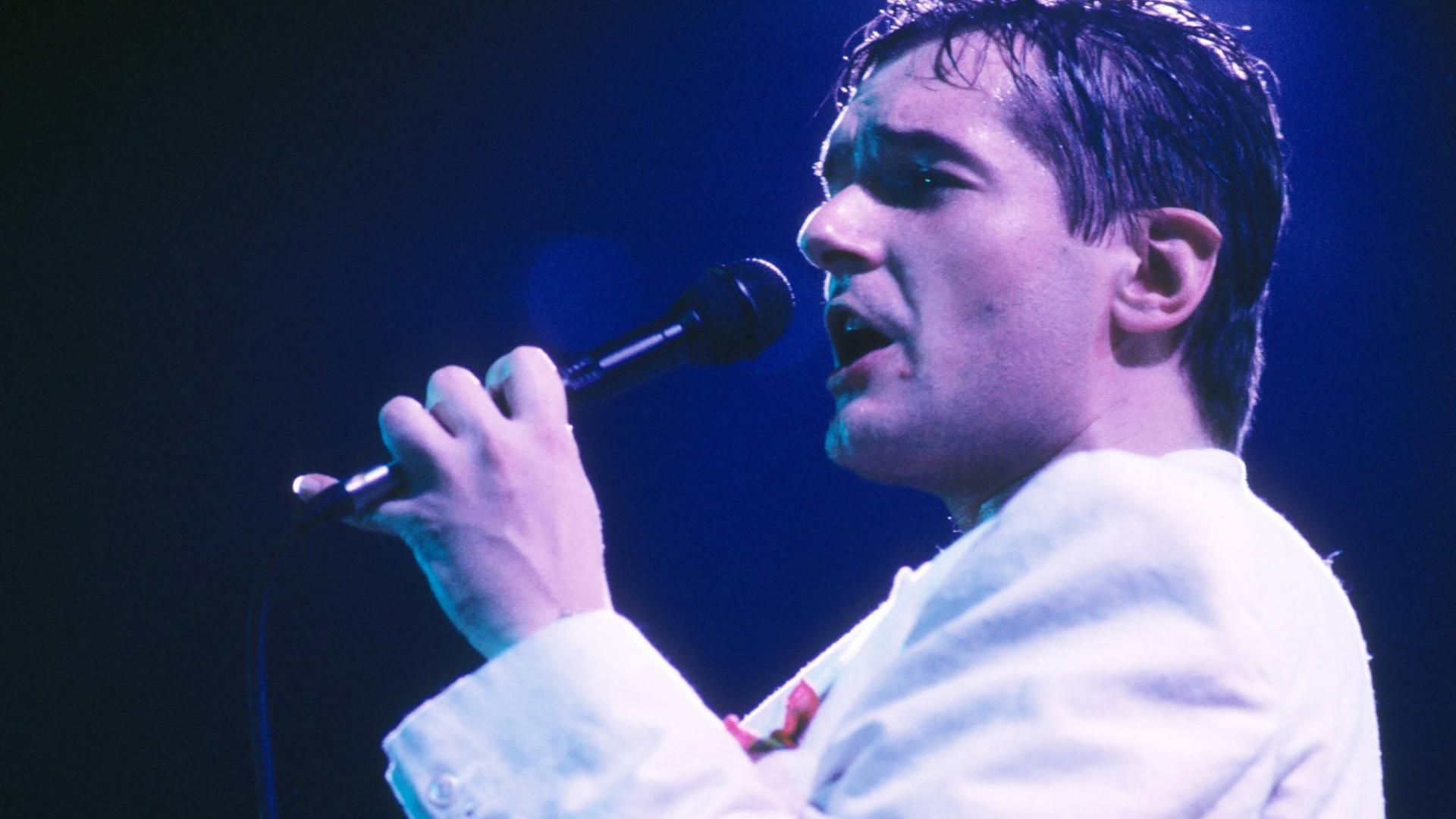 Der Popsänger Falco ist verschwitzt und singt in ein Mikrofon und trägt eine helle Jacke