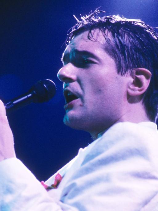 Der Popsänger Falco ist verschwitzt und singt in ein Mikrofon und trägt eine helle Jacke