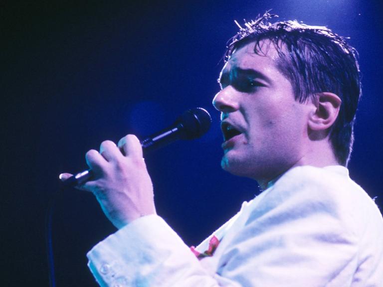 Der Popsänger Falco ist verschwitzt und singt in ein Mikrofon und trägt eine helle Jacke