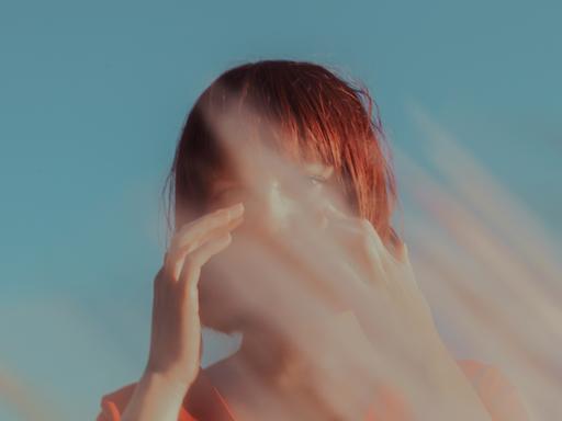 Eine junge Frau vor blauem Himmel. Vor ihrem Gesicht unscharfe rötlich-graue Streifen, wie bei Lichtstrahlen, Gräsern oder verwischter Farbe.