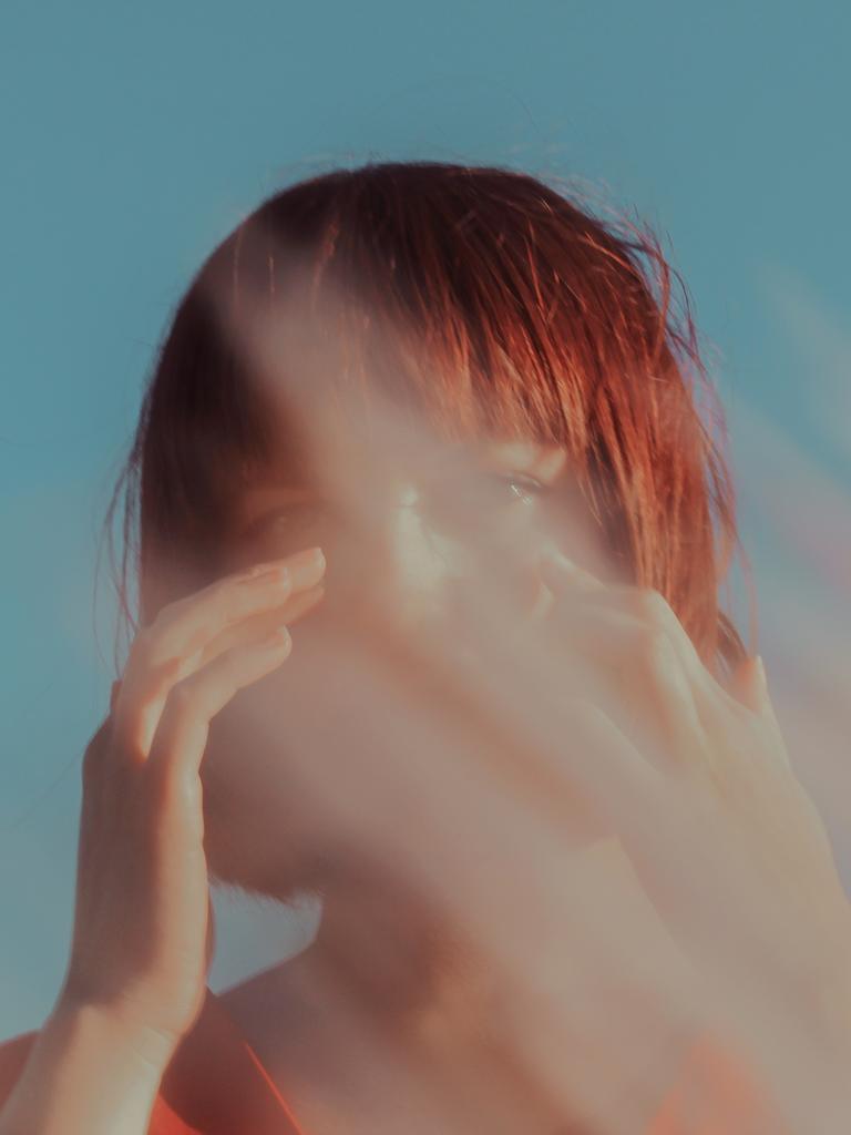 Eine junge Frau vor blauem Himmel. Vor ihrem Gesicht unscharfe rötlich-graue Streifen, wie bei Lichtstrahlen, Gräsern oder verwischter Farbe.