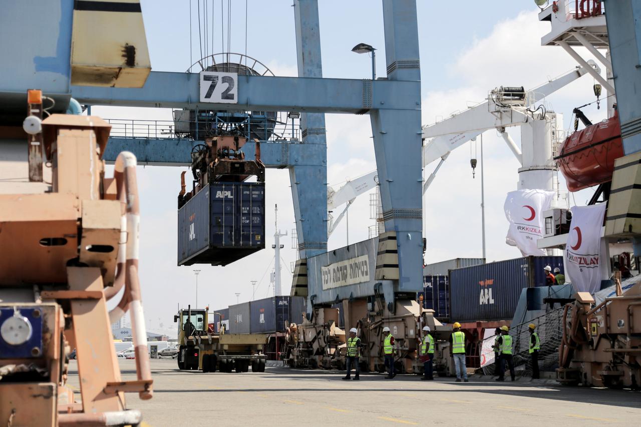 Der blauer Containerkran hebt gerade einen Container aus dem Schiff, das an einem Kai liegt. Davor Arbeiter in gelben Warnwesten und mit Helmen. 