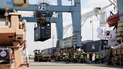 Der blauer Containerkran hebt gerade einen Container aus dem Schiff, das an einem Kai liegt. Davor Arbeiter in gelben Warnwesten und mit Helmen.