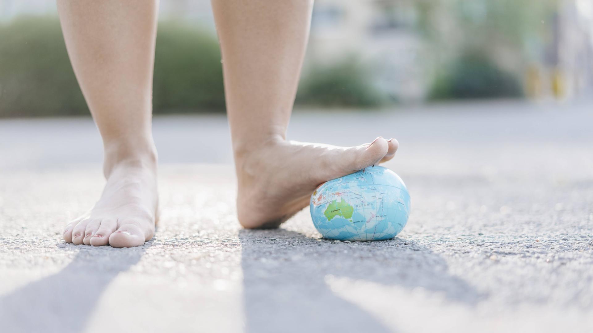 Zwei nackte Füße; der linkte drückt einen Erdkugel aus Stoff nach unten. Von hinten fällt Licht, sodass die Füße einen Schatten werfen.