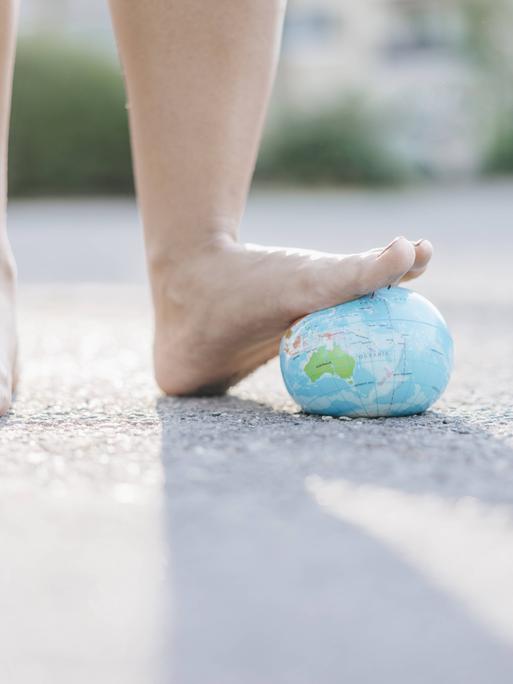 Zwei nackte Füße; der linkte drückt einen Erdkugel aus Stoff nach unten. Von hinten fällt Licht, sodass die Füße einen Schatten werfen. Zwei nackte Füße; der linkte drückt einen Erdkugel aus Stoff nach unten. Von hinten fällt Licht, sodass die Füße einen Schatten werfen.