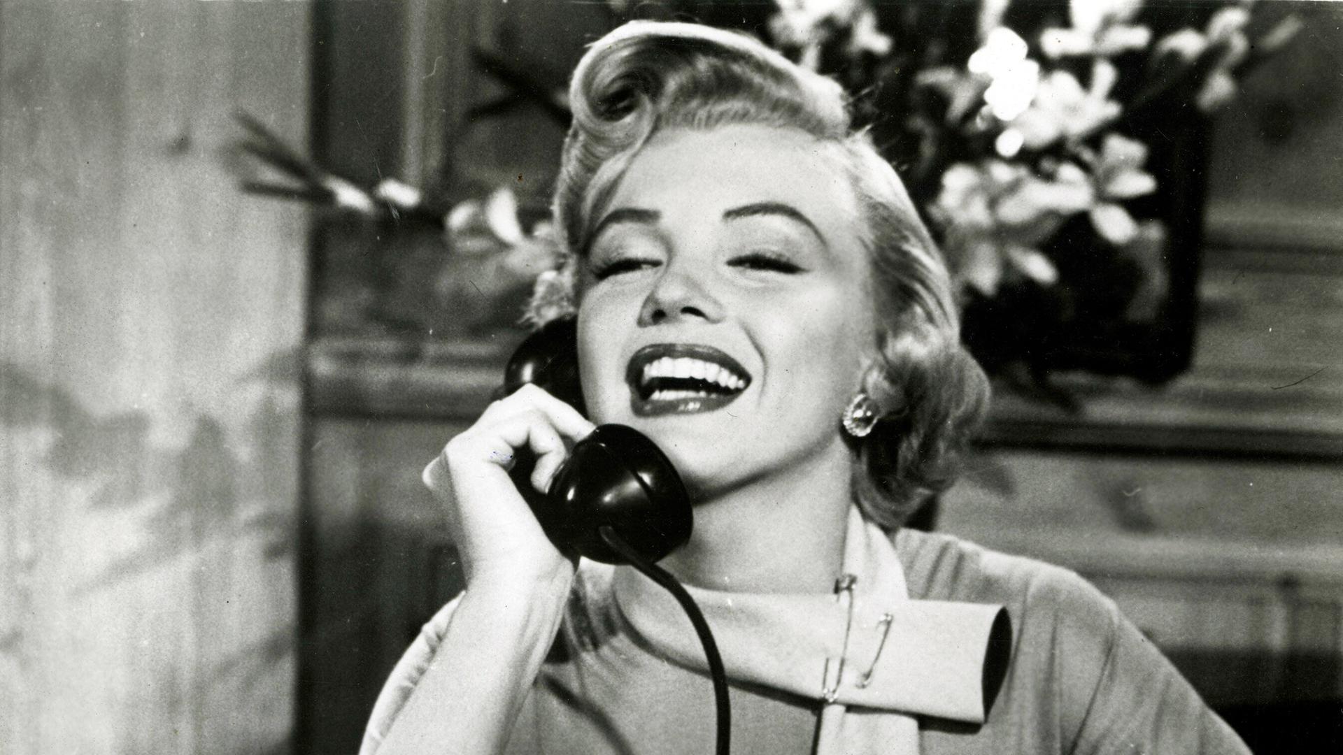 Marilyn Monroe mit platinblonder Föhnfrisur sitzt  im Film "Liebling, ich werde jünger" an einem Schreibtisch hält einen Telefonhörer an ihr rechtes Ohr und lächelt breit.