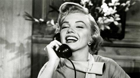 Marilyn Monroe mit platinblonder Föhnfrisur sitzt  im Film "Liebling, ich werde jünger" an einem Schreibtisch hält einen Telefonhörer an ihr rechtes Ohr und lächelt breit.