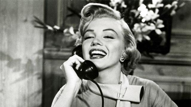 Marilyn Monroe mit platinblonder Föhnfrisur sitzt  im Film "Liebling, ich werde jünger" an einem Schreibtisch hält einen Telefonhörer an ihr rechtes Ohr und lächelt breit.
