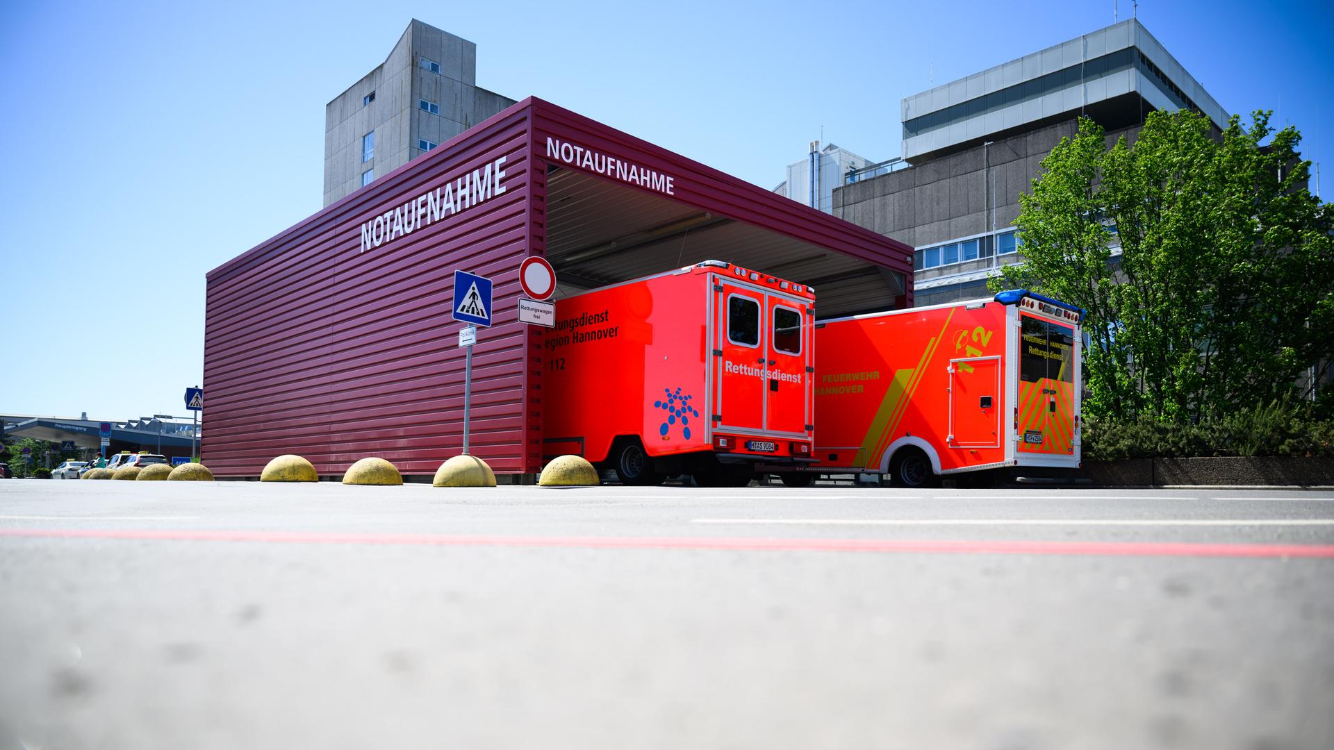 Zwei Rettungswagen stehen an einer Notaufnahme. Das Gebäude ist weinrot, im Hintegrund ist ein Krankenhaus zu sehen.