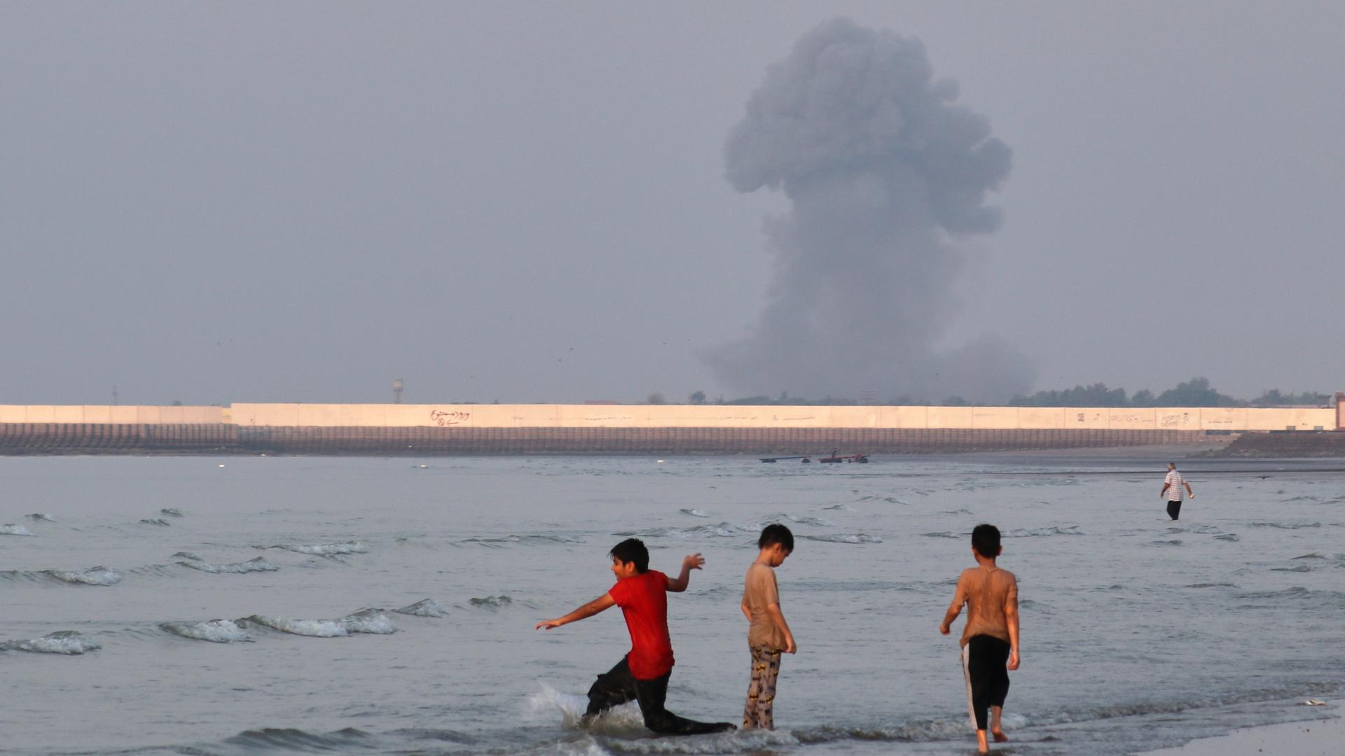Iran, Bandar Abbas: Drei Jungen spielen im seichten Wasser der Straße von Hormus, während im Hintergrund vor der Küste von Bandar Abbas im Iran eine Rauchwolke nach einer Explosion aufsteigt. Iran, Bandar Abbas: Drei Jungen spielen im seichten Wasser der Straße von Hormus, während im Hintergrund vor der Küste von Bandar Abbas im Iran eine Rauchwolke nach einer Explosion aufsteigt.