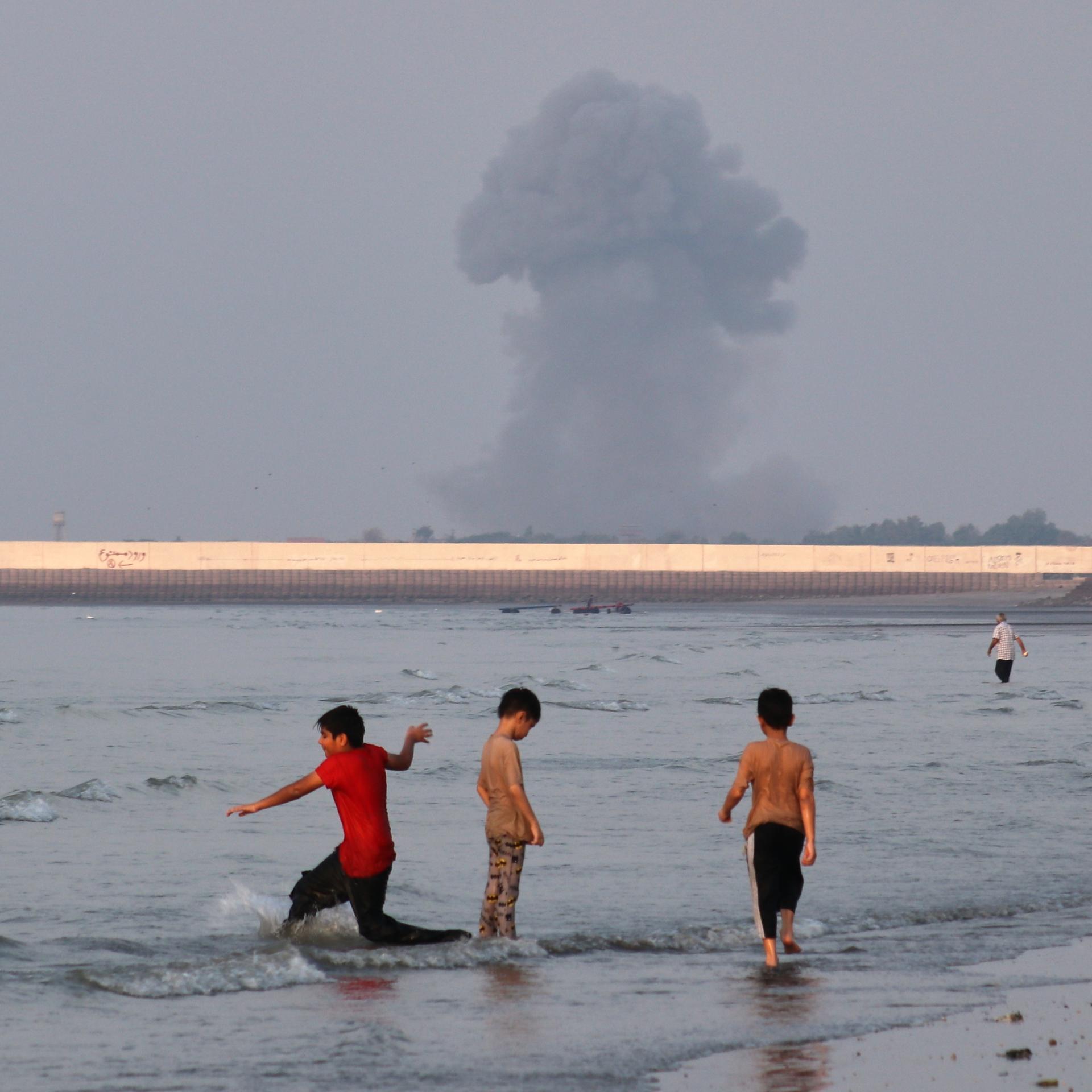 Iran, Bandar Abbas: Drei Jungen spielen im seichten Wasser der Straße von Hormus, während im Hintergrund vor der Küste von Bandar Abbas im Iran eine Rauchwolke nach einer Explosion aufsteigt.