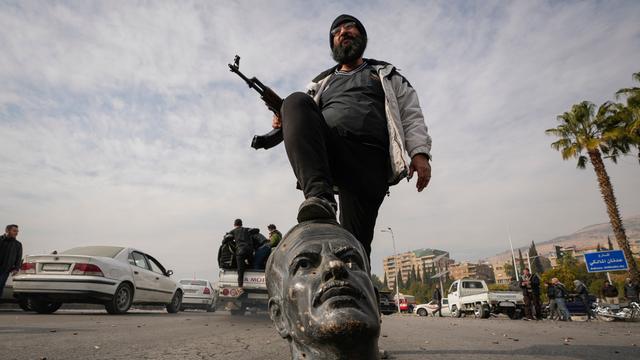 Ein Kämpfer mit Maschinen-Gewehr in der Hand tritt auf eine zerbrochene Statue von dem verstorbenen syrischen Präsidenten Hafez Assad. Das war der Vater von Baschar al-Assad. 