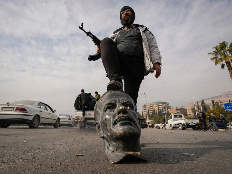 Ein Oppositionskämpfer mit Maschinengewehr in der Hand tritt am Sonntag, 8. Dezember 2024, in Damaskus, Syrien, auf eine zerbrochene Büste des verstorbenen syrischen Präsidenten Hafez Assad. 