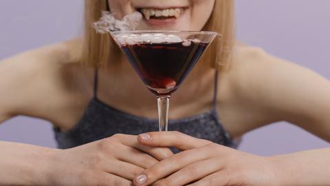 Eine Frau mit Vampirzähnen sitzt vor einem Cocktailglas mit roter Flüssigkeit.