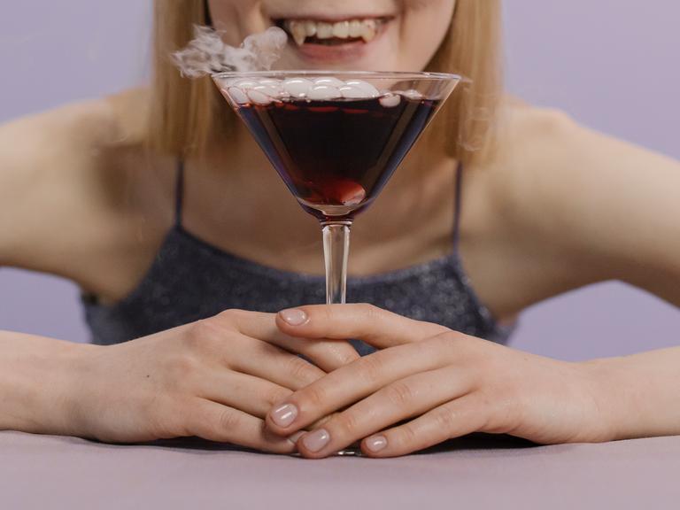 Eine Frau mit Vampirzähnen sitzt vor einem Cocktailglas mit roter Flüssigkeit.