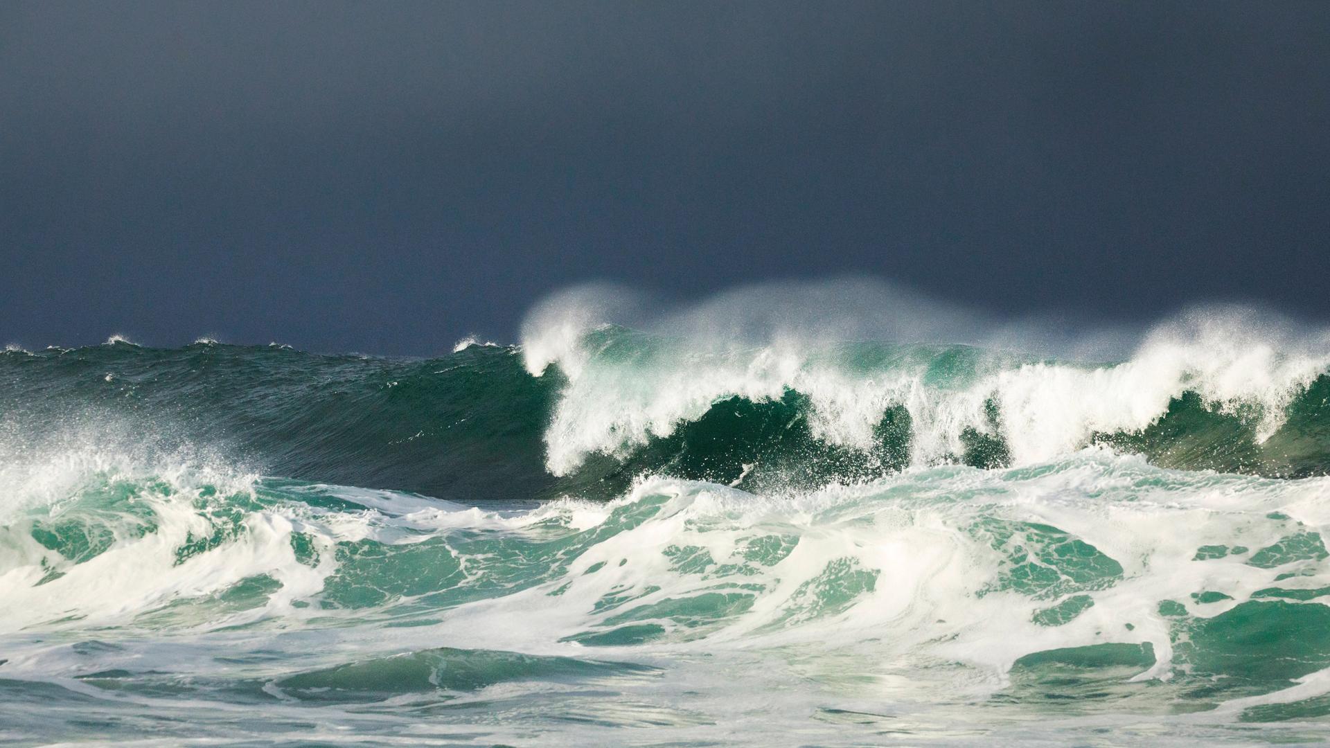 Aufnahme einer großen Welle, die bricht. Über dem Meer sieht man einen grauen Himmel.