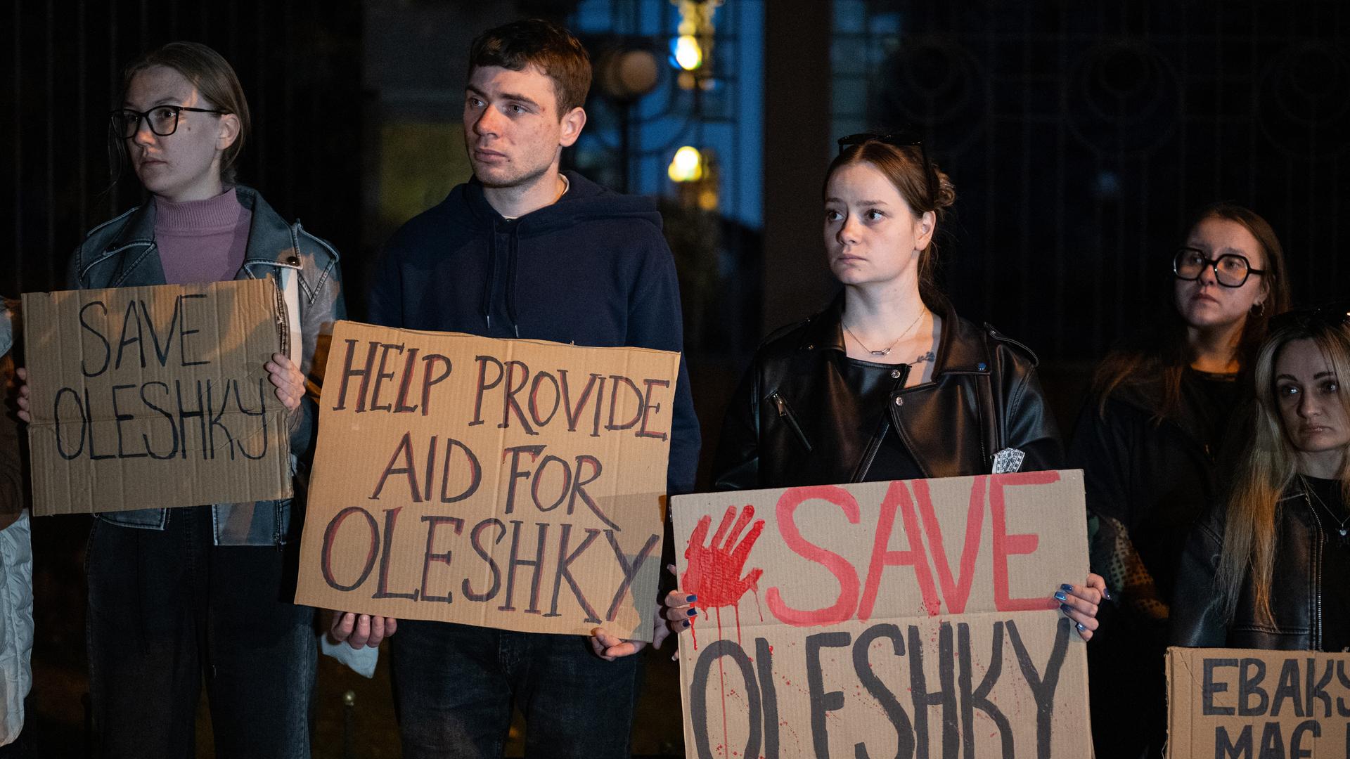 Mehrere Männer und Frauen halten Pappschilder, auf denen unter anderem "Save Oleshky" steht. Es ist dunkel. 