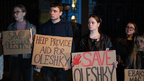 Mehrere Männer und Frauen halten Pappschilder, auf denen unter anderem "Save Oleshky" steht. Es ist dunkel. 