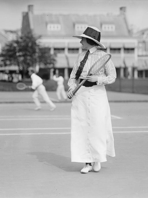 Eine historische Schwarz-Weiß-Fototrafie zeigt die Tennisspielerin Miss Frances Lippett mit Krawatte, Hut und Tennisschläger bei einem Turnier um 1913. 