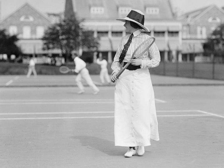 Eine historische Schwarz-Weiß-Fototrafie zeigt die Tennisspielerin Miss Frances Lippett mit Krawatte, Hut und Tennisschläger bei einem Turnier um 1913. 