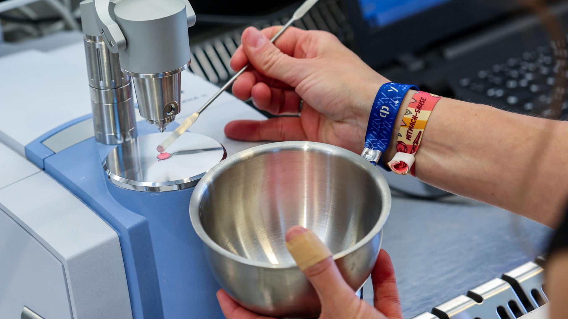 Man sieht die Hände einer Person, die mit links eine metallene Schüssel hält und mit rechts mit einer Pipette eine Flüssigkeit auf eine Fläche streicht.