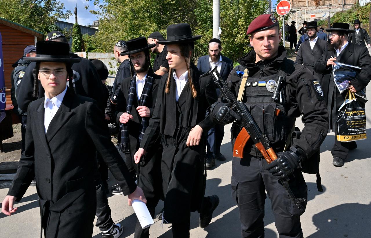 Ein bewaffneter Polizist bewacht jüdische Pilger in traditionellem Gewand.