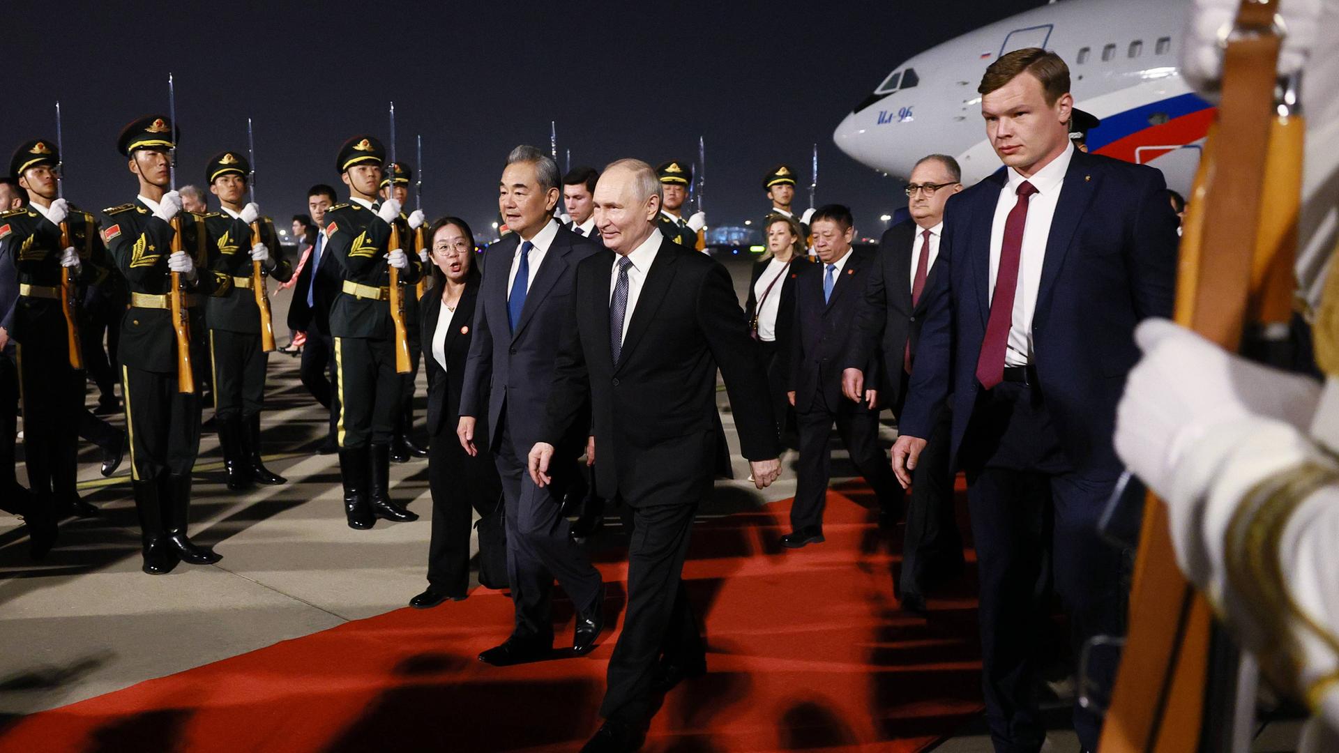 Der russische Präsident Wladimir Putin läuft am Flughafen in Peking über einen roten Teppich. Neben ihm laufen der chinesische Außenminister Wang Yi sowie mehrere Begleiter. Im Hintergrund stehen chinesische Soldaten in Uniform.