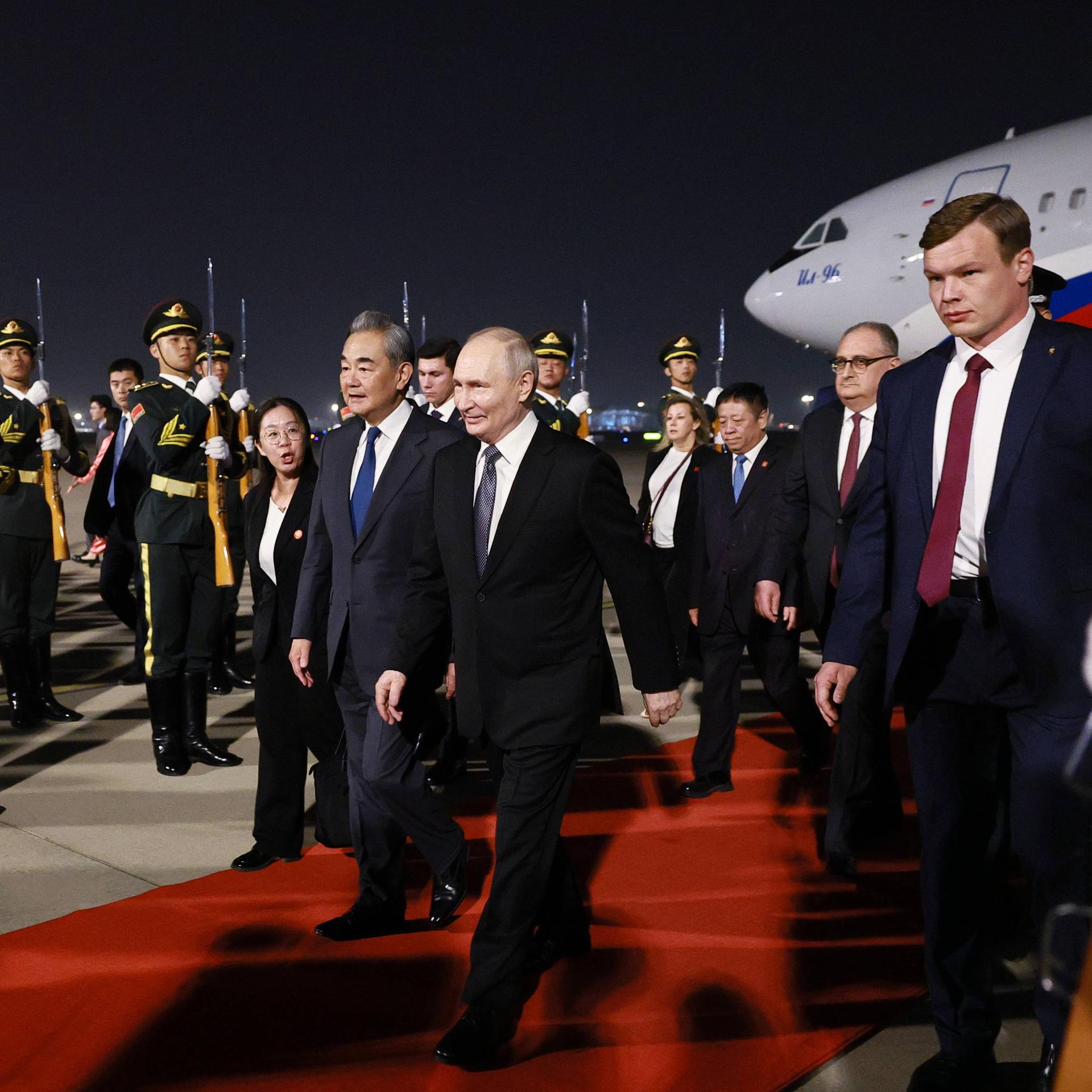 Der russische Präsident Wladimir Putin läuft am Flughafen in Peking über einen roten Teppich. Neben ihm laufen der chinesische Außenminister Wang Yi sowie mehrere Begleiter. Im Hintergrund stehen chinesische Soldaten in Uniform.