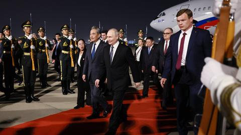 Der russische Präsident Wladimir Putin läuft am Flughafen in Peking über einen roten Teppich. Neben ihm laufen der chinesische Außenminister Wang Yi sowie mehrere Begleiter. Im Hintergrund stehen chinesische Soldaten in Uniform.